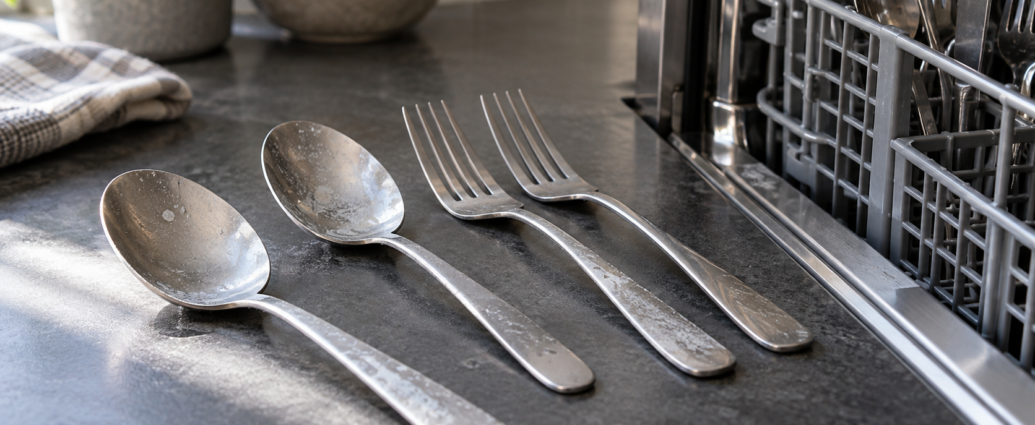 Cucchiai e forchette in acciaio con una leggera patina bianca appoggiati accanto al cestello della lavastoviglie